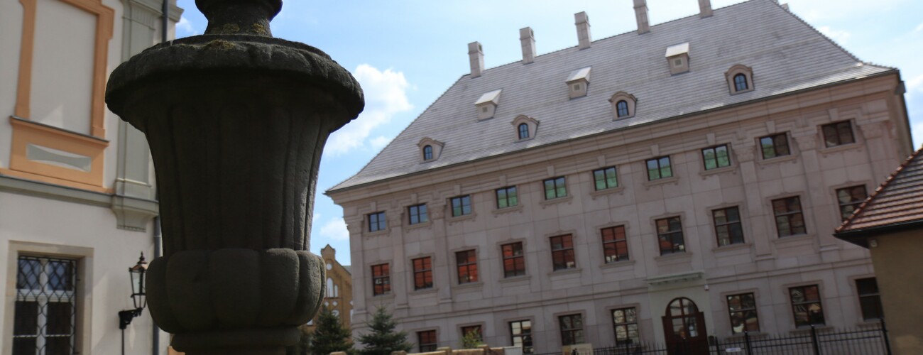 Zdjęcie frontu biblioteki wraz z kamienną rzeźbą stojącą na pagórku przed biblioteką.